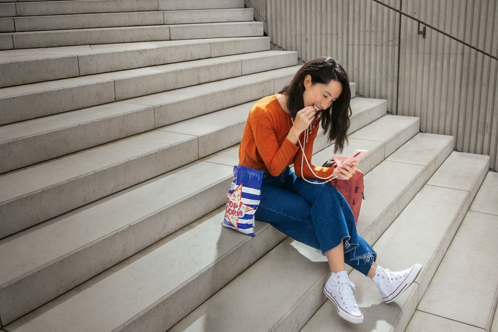 Eine junge Frau sitzt auf einer Beton-Treppe, isst Popcorn und schaut dabei auf ihr Smartphone.