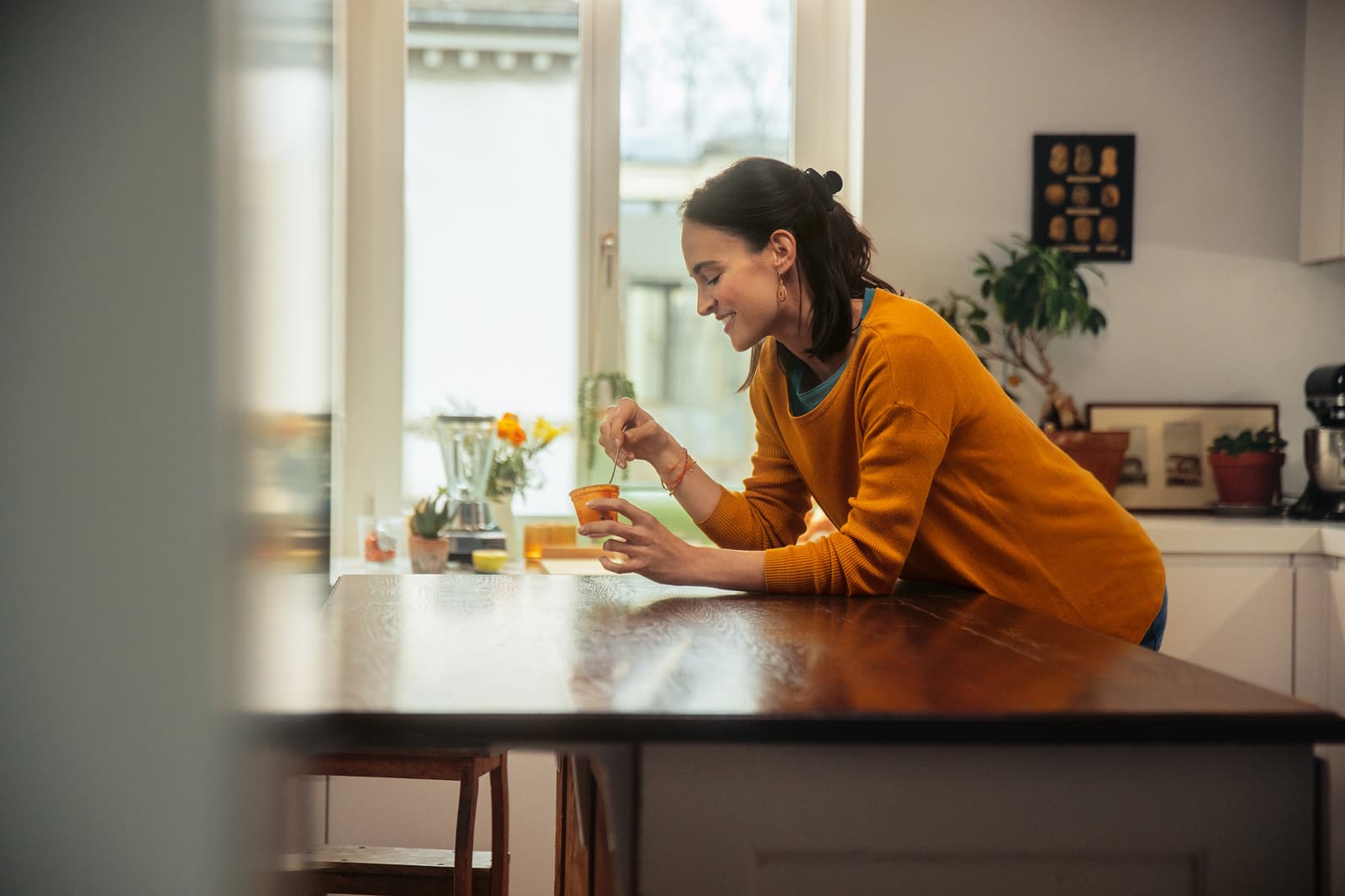 Una signora si appoggia al tavolo della cucina mentre assaggia uno yogurt.